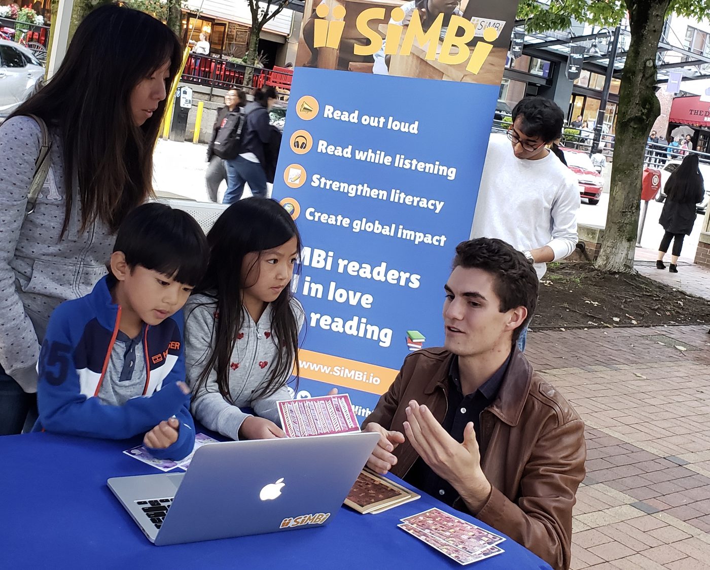 Alex at a street booth showing two children a laptop